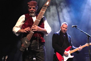 Stickist on stage with guitarist in background, blue wash background with white rays through smoke.